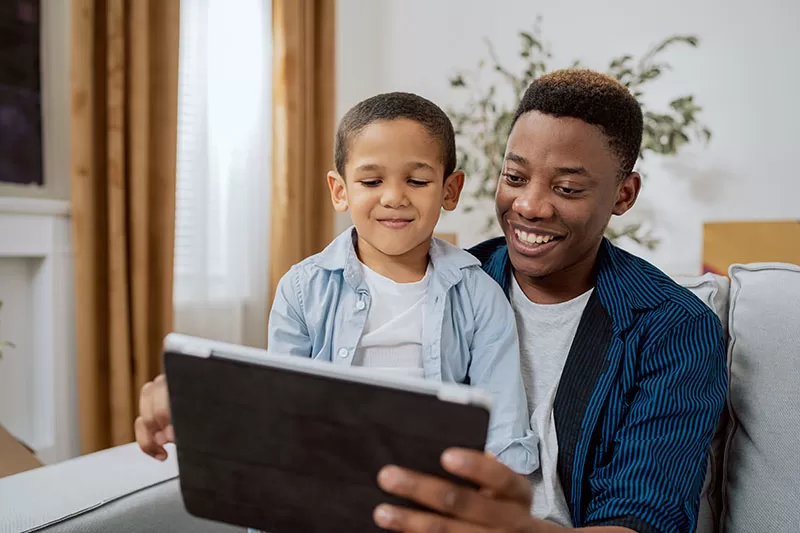 Proud father sits on living room couch holding handsome young son on lap, tablet in hand, they are having a video conversation via webcam